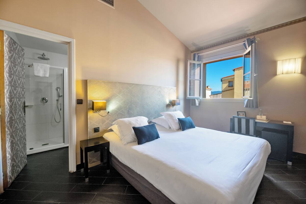 A hotel room with a large bed, a bedside table, a nightstand, a mirror, a window with a view, and a shower area.