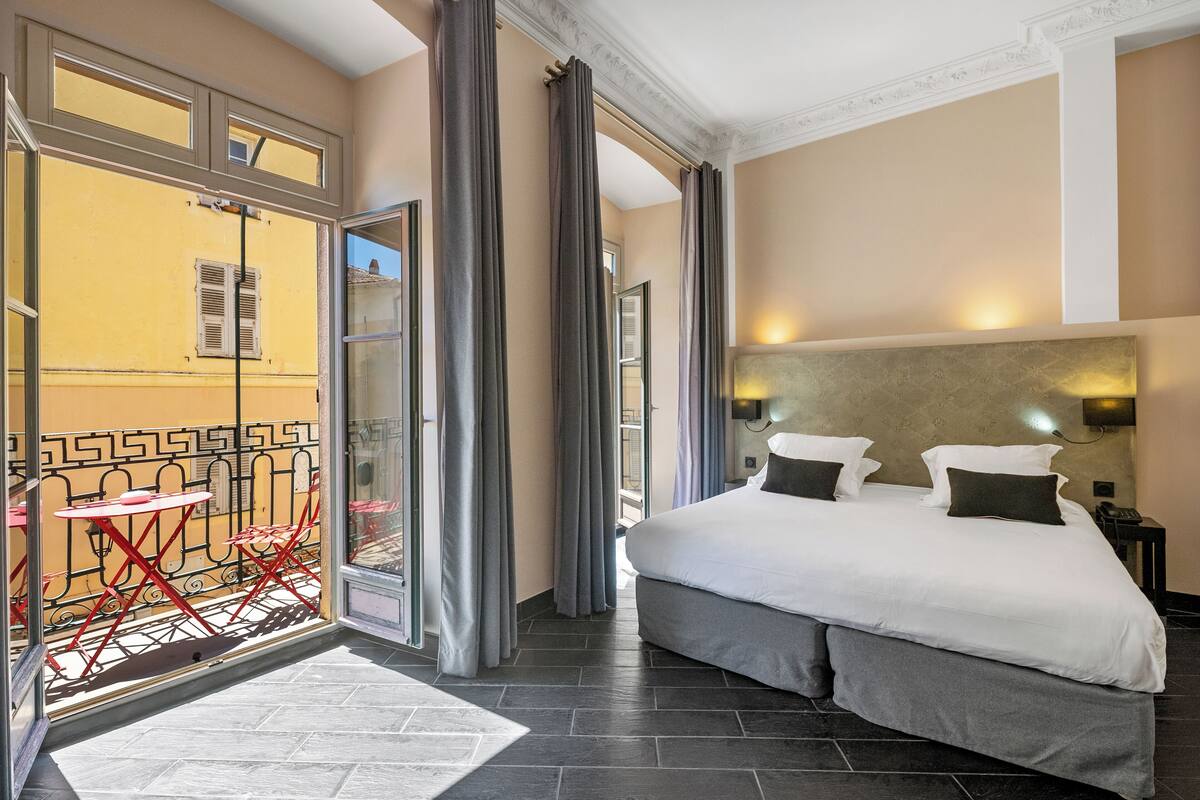 A hotel room with a large bed, a small table, and a balcony with red chairs.