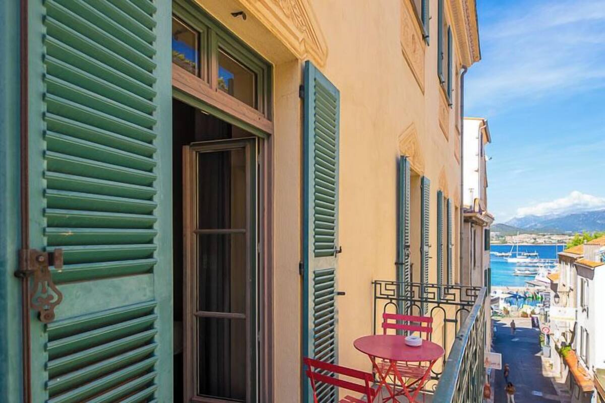 A narrow street with a building featuring green shutters and a balcony with a red table and chair.