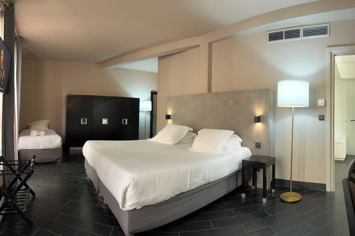 A modern hotel room with a large bed, a desk, and a chair. There is a wardrobe, a bedside table, and a lamp.