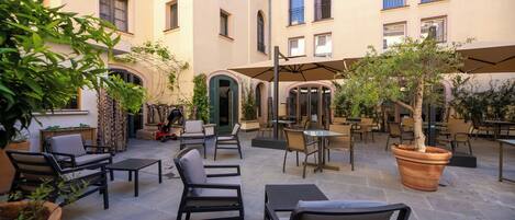 An outdoor patio area with multiple tables and chairs, surrounded by potted plants and a building with multiple windows and balconies.