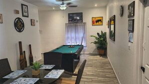 Ein Billardtisch mit Spielstöcken, ein Esstisch mit Stühlen und ein Deckenventilator in einem Raum mit Holzboden und Wanddekorationen.