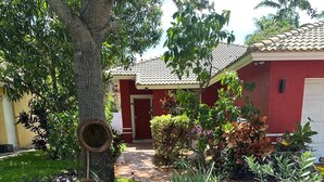 Ein Garten mit Baum, verschiedenen Pflanzen, einem roten Gebäude mit Ziegeldach und einem Weg, der zu einer Tür führt.