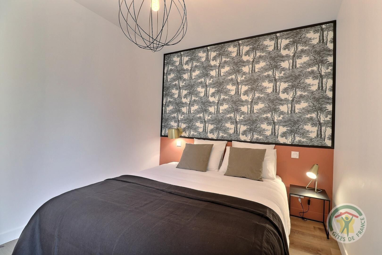Une chambre à coucher avec un grand lit, un tableau représentant des arbres accroché au mur, des lampes de chevet et un luminaire au plafond.