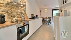 A modern kitchen with a stone wall, a coffee machine, a built-in oven, and a dining area with a table and chairs. - Victor - RIVES DU COUESNON (RIVES-DU-COUESNON)