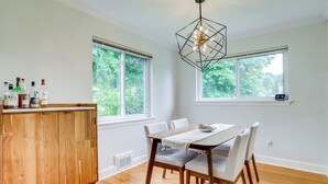A dining area with a wooden table, white chairs, and a pendant light. There are windows with a view of greenery outside. - Cozy Modern Single Family 5BD 2BA Home (Falls Church)