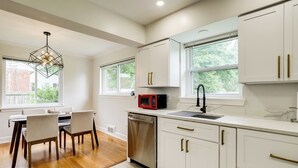A modern kitchen with white cabinets, a stainless steel dishwasher, a red microwave, and a black faucet. There is a dining table with chairs and a pendant light above it. - Cozy Modern Single Family 5BD 2BA Home (Falls Church)