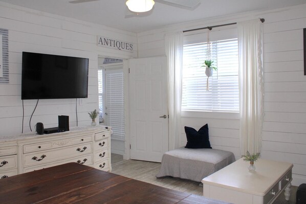 A room with a wooden floor, a white dresser with drawers, a flat-screen TV mounted on the wall, a small bench with a cushion, and a window with blinds and a hanging plant.