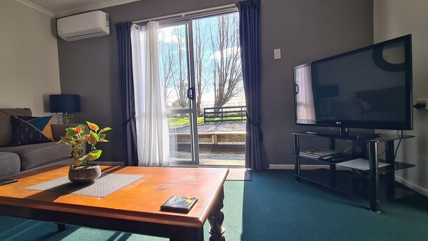 A living room with a sofa, a coffee table with a plant, a TV on a stand, and a sliding glass door leading outside. - Kawakawa Cottage - lovely rural outlook. Convenient location to Manfield Park  (Feilding)