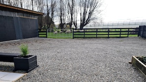 Un patio de grava con una jardinera negra, una cerca de metal y vistas a una zona verde con árboles y un invernadero al fondo.