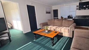 A living room with a sofa, a coffee table, and a TV. There is a kitchen area with cabinets and a sink. - Kawakawa Cottage - lovely rural outlook. Convenient location to Manfield Park  (Feilding)