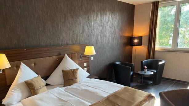 A hotel room with a large bed, two bedside lamps, a chair, and a small table. - Hotel Pasa Paradies (Hanau)