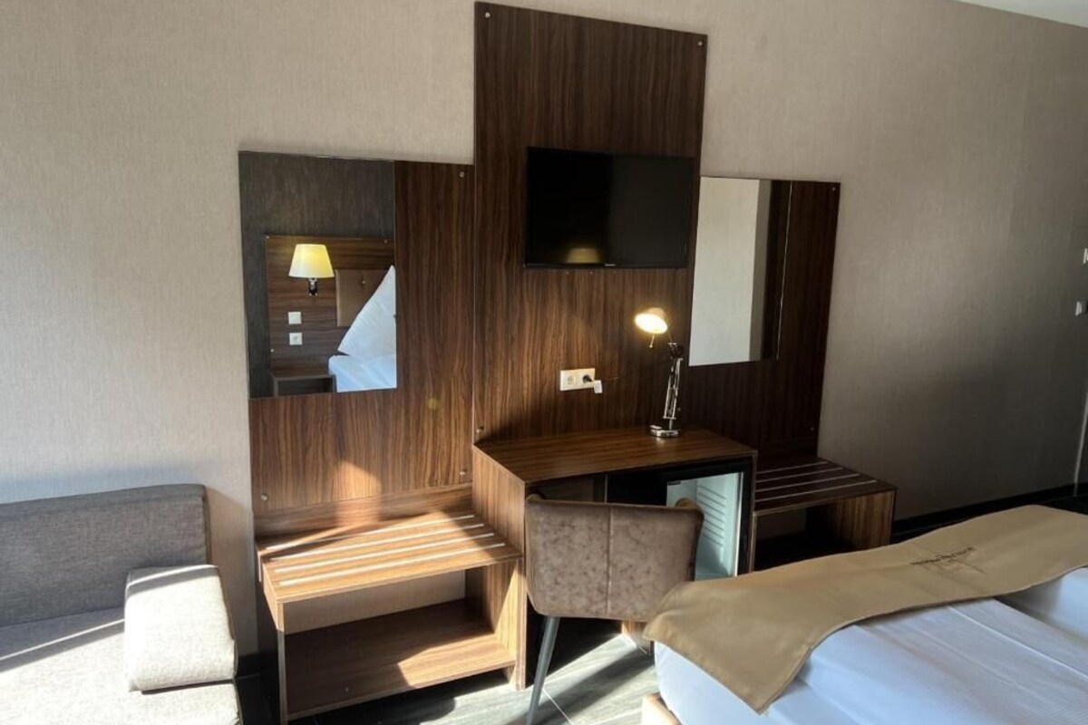 A hotel room with a bed, a desk, a chair, a TV mounted on the wall, and a lamp.