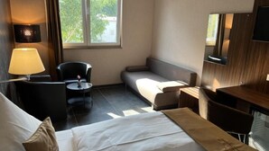 A hotel room with a bed, a desk, a chair, a sofa, a lamp, and a TV. - Hotel Pasa Paradies (Hanau)