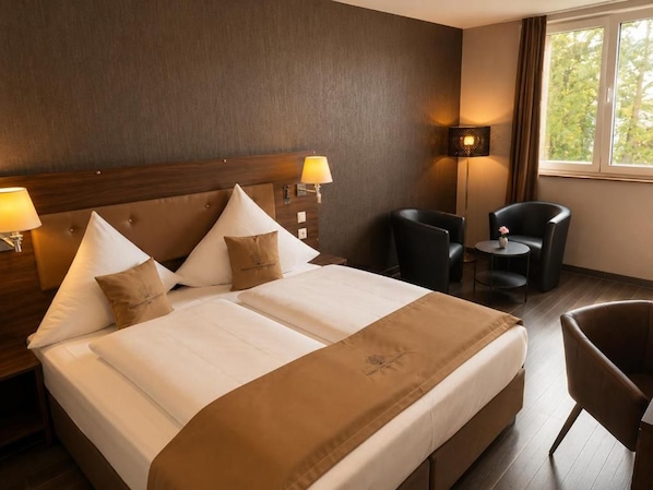 Quarto de hotel com uma cama grande, duas luminárias de cabeceira, uma cadeira, uma mesa pequena e uma janela com cortinas.