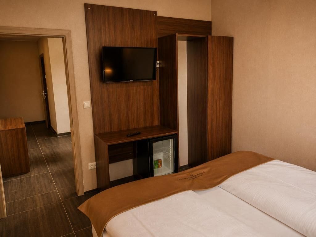 A hotel room with a bed, a television mounted on a wooden panel, a small refrigerator, and a wardrobe.
