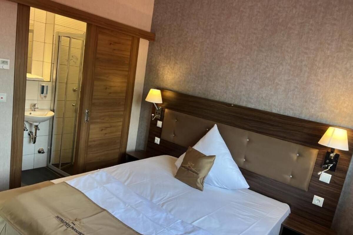 Hotellihuone, jossa on sänky, kaksi yöpöytälamppua, puinen sängynpääty ja kylpyhuone, joka näkyy avoimesta ovesta.