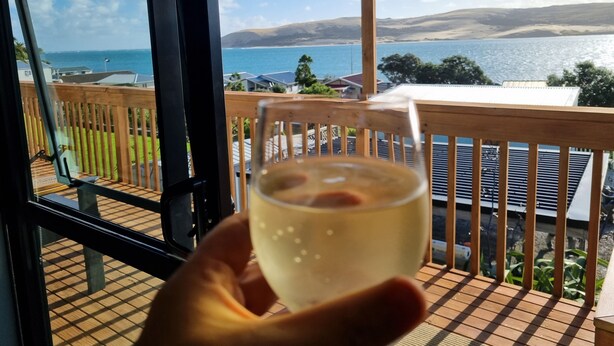 A hand holding a glass of white wine on a wooden deck with a view of a lake and mountains.