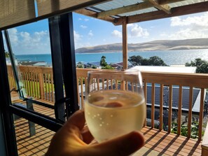 A hand holding a glass of white wine on a wooden deck with a view of a lake and mountains. - Gorgeous cottage, stunning ocean views,  1 min to the beach! Relax & recharge 😍 (Opononi)