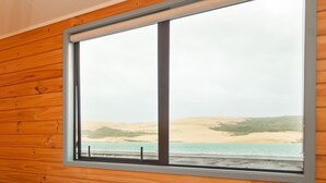 A large window with a metal frame, wooden wall paneling, and a view of a beach with sand dunes and water.