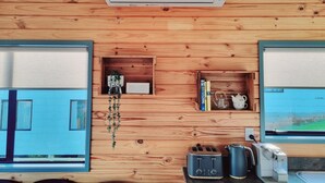 A cabin interior with wooden walls, a built-in air conditioner, a small shelf with books and a kettle, a toaster, and a kettle on the counter.