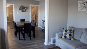 A modern living room with a grey sofa, a dining area with a wooden table and chairs, and a hallway leading to a kitchen.