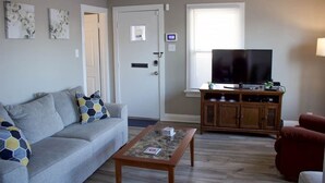 A living room with a grey sofa, a coffee table, a TV on a wooden cabinet, and a door with a security panel.