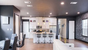 A modern kitchen with white cabinets, a central island with bar stools, and a dining area with a white table and chairs. - Overnight Gallery • The Art House • Near Downtown Dallas • Maduro Collection (Dallas)