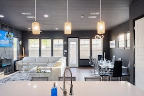 A modern kitchen with a central island, dining area with a long table and chairs, a sofa, and pendant lighting. - Overnight Gallery • The Art House • Near Downtown Dallas • Maduro Collection (Dallas)