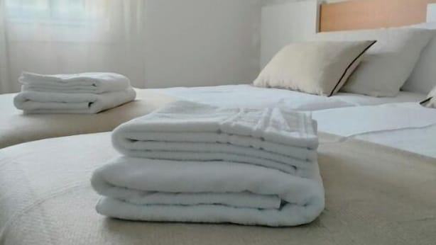 Ein ordentlich bezogenes Bett mit gefalteten weißen Handtüchern, einem hölzernen Kopfteil und einem Fenster mit transparenten Vorhängen.