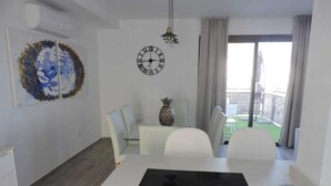 Una cucina moderna con isola centrale, zona pranzo con sedie bianche, un orologio al muro e vista verso il balcone esterno.