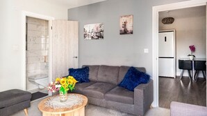 Ein modernes Wohnzimmer mit grauem Sofa, blauen Kissen, einem runden Holztisch mit Blumenvase und einem Essbereich mit Tisch und Stühlen.