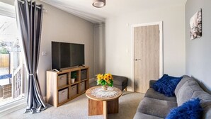 Ein Wohnzimmer mit grauem Sofa, einem runden Holztisch mit einer Vase voller Sonnenblumen, einem Flachbildfernseher auf einem Holzständer und einer Tür mit heller Holzvertäfelung.