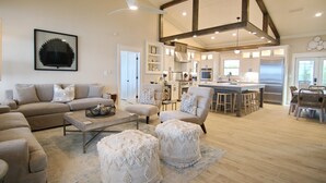 A spacious living room with a sofa set, ottomans, and a coffee table. A kitchen with modern appliances and bar stools is visible in the background. - Bayfront, Pool, Pier, 2 Hot Tubs!, Sandy Beach, Private, steps from beachfront (Saint George Island)