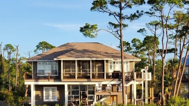 A two-story house with a wooden deck leading to the entrance, surrounded by water and trees. - Bayfront, Pool, Pier, 2 Hot Tubs!, Sandy Beach, Private, steps from beachfront (Saint George Island)