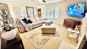 Ein Wohnzimmer mit einem Ecksofa, einem Hocker und einem an der Wand befestigten Fernseher.