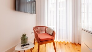 A modern living room with a wooden floor, a red cushioned chair, a small table with a plant and remote control, and a large TV on the wall. - S. Bento Apart IV (G) (Oporto)