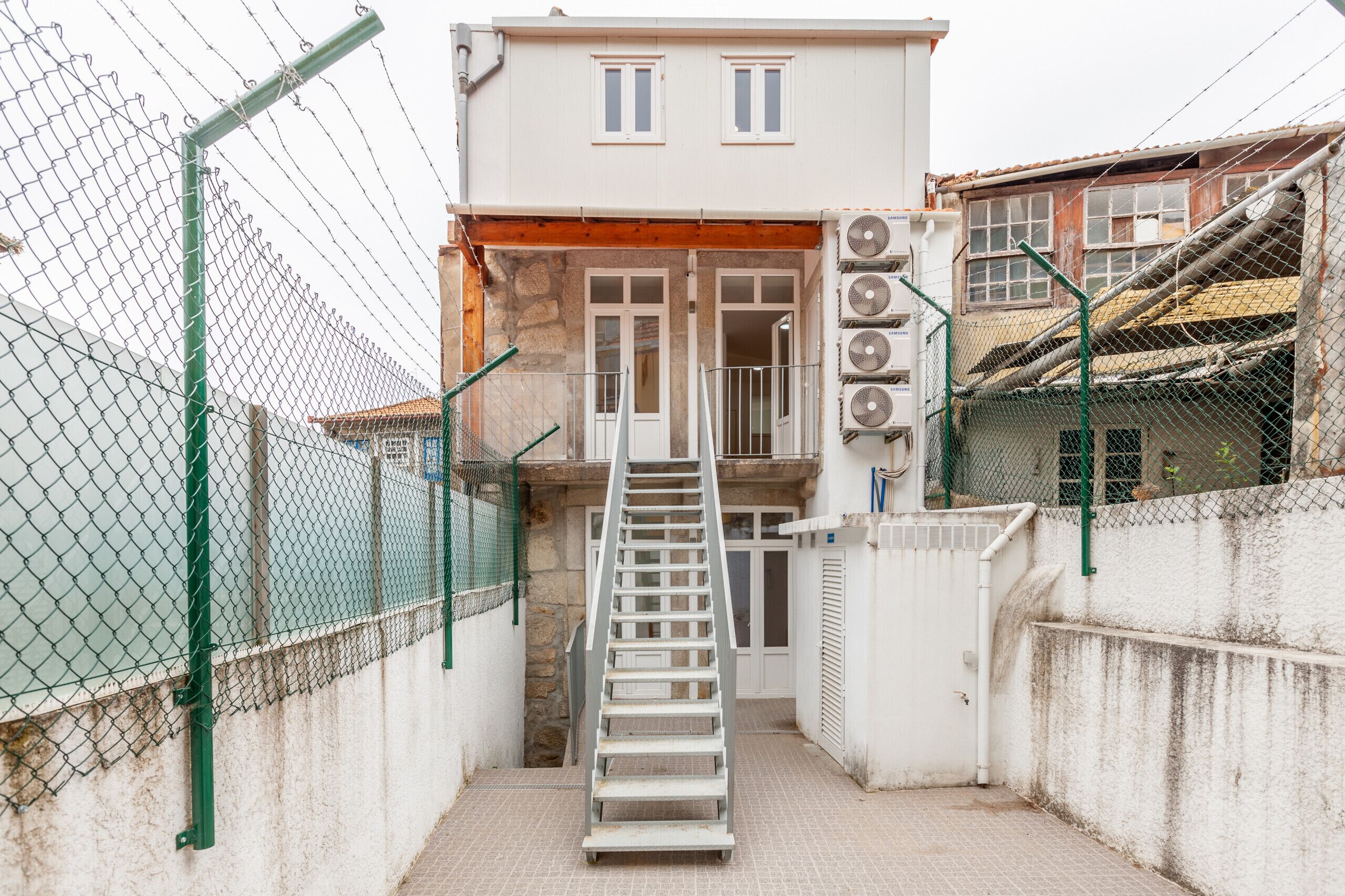 A building with a staircase leading to a door, surrounded by chain-link fences and a concrete wall.