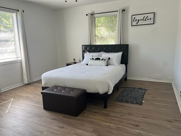 A bedroom with a bed, a nightstand, a small table with a plant, a black ottoman, a black and white rug, and a window with curtains.