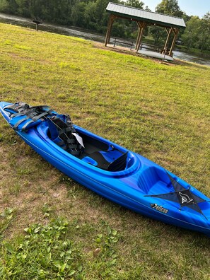 A blue kayak with a brand name and a paddle, placed on grass near a river and a gazebo.