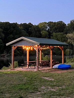 Un pabellón de madera con luces de cuerda, mesas de picnic y un inflable azul en un espacio cubierto de césped cerca del agua.