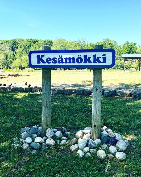 Un letrero con el nombre 'Kesämäki' en una tipografía clara y audaz, sujeto a dos postes de madera con una base de piedras redondeadas.