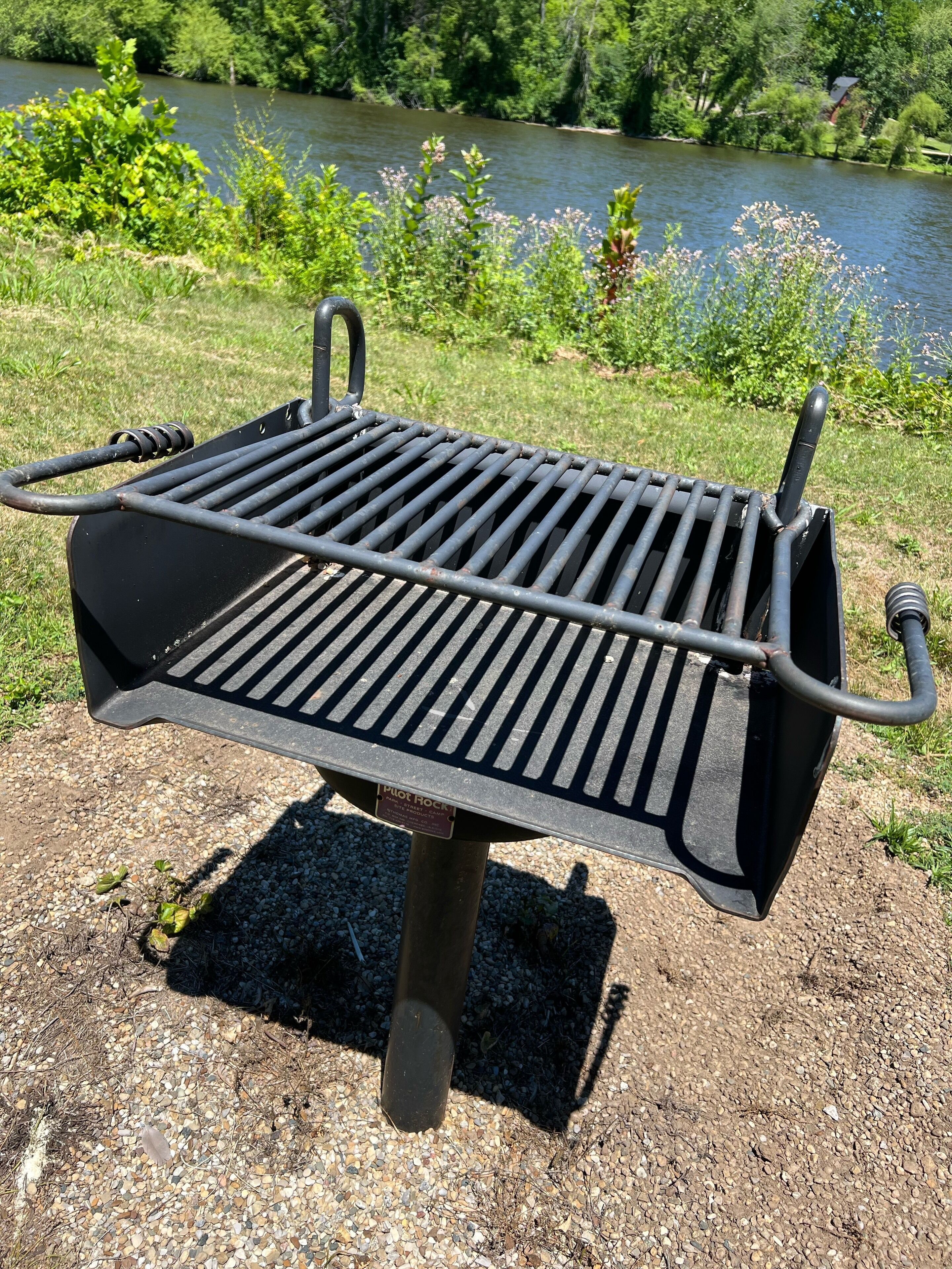 Un barbecue portable doté d’une grille à fentes, placé sur une surface de gravier, avec une rivière et de la végétation en arrière-plan.