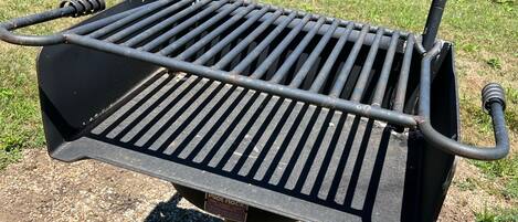 Un barbecue portable doté d’une grille à fentes, placé sur une surface de gravier, avec une rivière et de la végétation en arrière-plan.