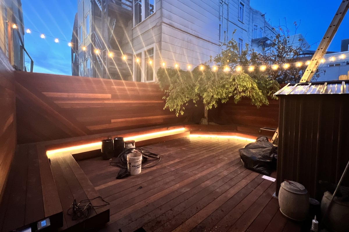 Une terrasse en bois, décorée de guirlandes lumineuses, avec un arbre et un bâtiment en arrière-plan.