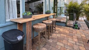 Een buitenpatio met een houten bar en krukken, een afvalbak en potplanten.