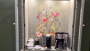 Een koffiehoek in vintage stijl, met een koffiezetapparaat, kopjes en een decoratief paneel met vogels en bloemen.