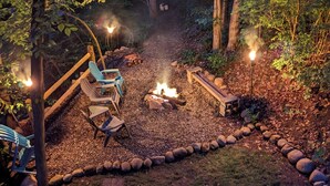 A forested backyard with a fire pit, seating area, and a path leading to a wooden deck.