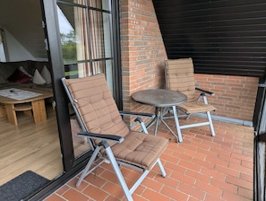 A patio with two cushioned chairs and a small round table, a wooden bench, and a glass door leading inside.