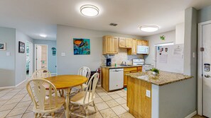 A compact kitchen with wooden cabinets, a white refrigerator, a microwave, and a coffee maker. There is a dining table with chairs and a colorful painting on the wall.